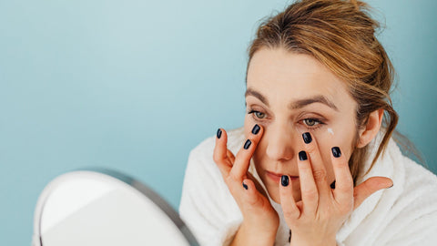 Woman gently applying a small amount of eye cream beneath her eye in a mirror, natural skincare photography. Photo via Pexels.